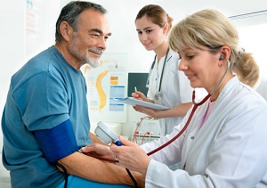 This is a picture of a female Doctor taking a male patient's pulse and blood pressure. There is a female Nurse in the background holding a Note pad and pen. they are all smiling.